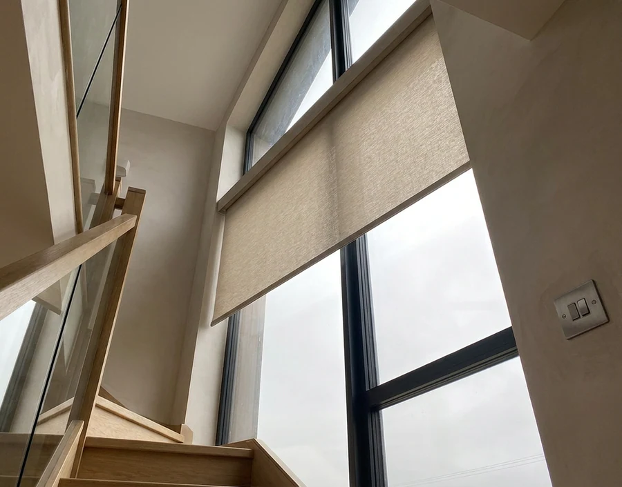 Motorised roller blind lowered over a tall stairwell window, a small wall-mounted control switch beside the frame