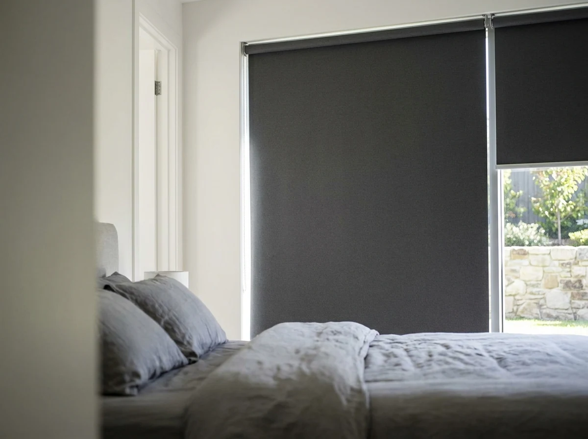 Estate-home bedroom in Sunninghill with a blockout roller blind lowered over a tall window, side angle showing the headrail and crisp daylight along the edge